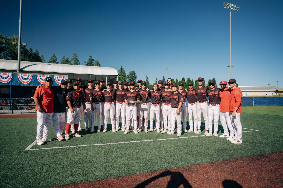 The Cardinals team lined up together in black jerseys and pinstripe pants, with coaches in red at each end, on a turf field under a blue sky.