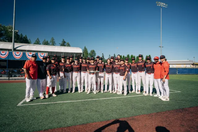 The Cardinals team lined up together in black jerseys and pinstripe pants, with coaches in red at each end, on a turf field under a blue sky.