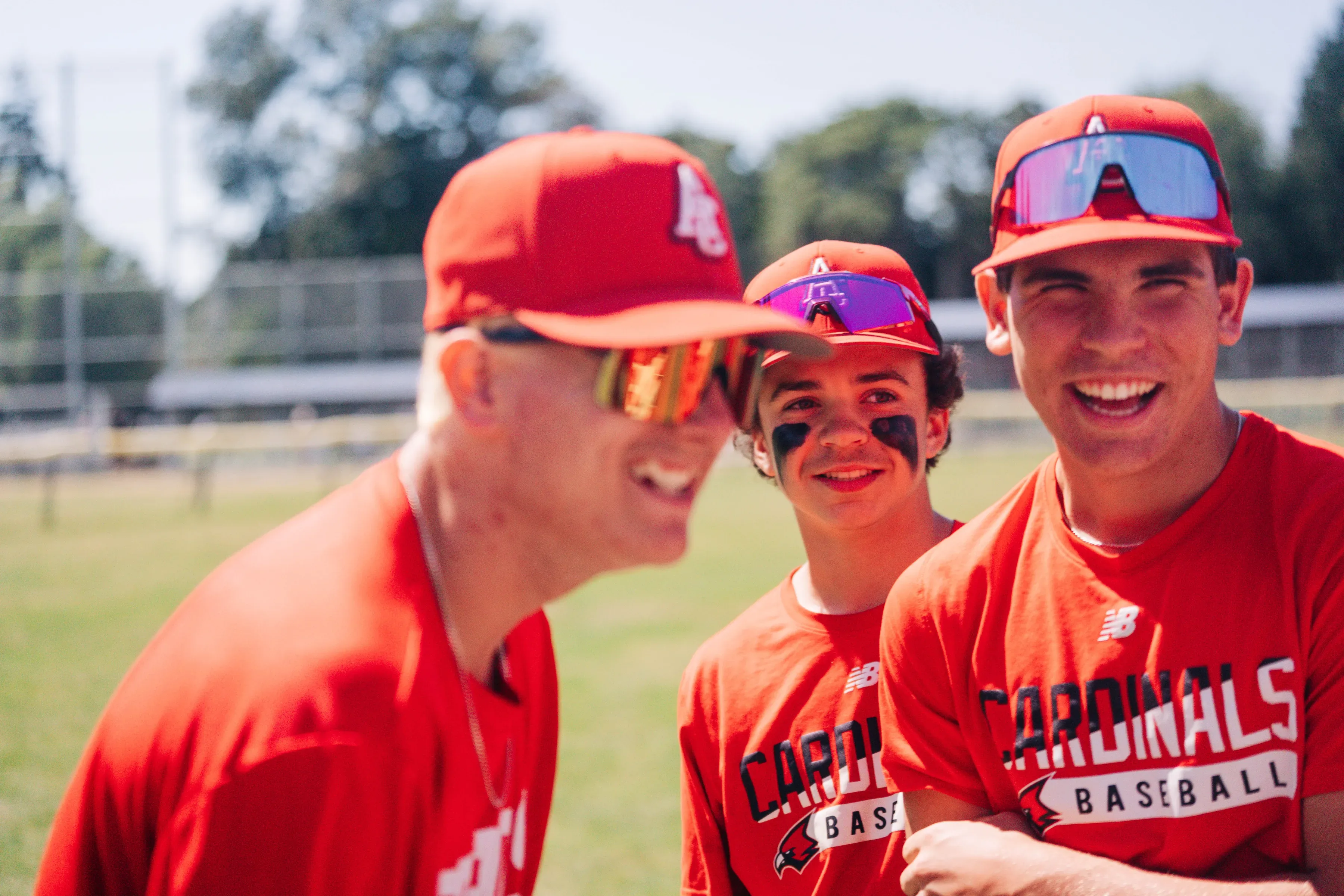 Three Cardinals players in red Cardinals Baseball shirts and red caps, smiling and laughing together on the field.