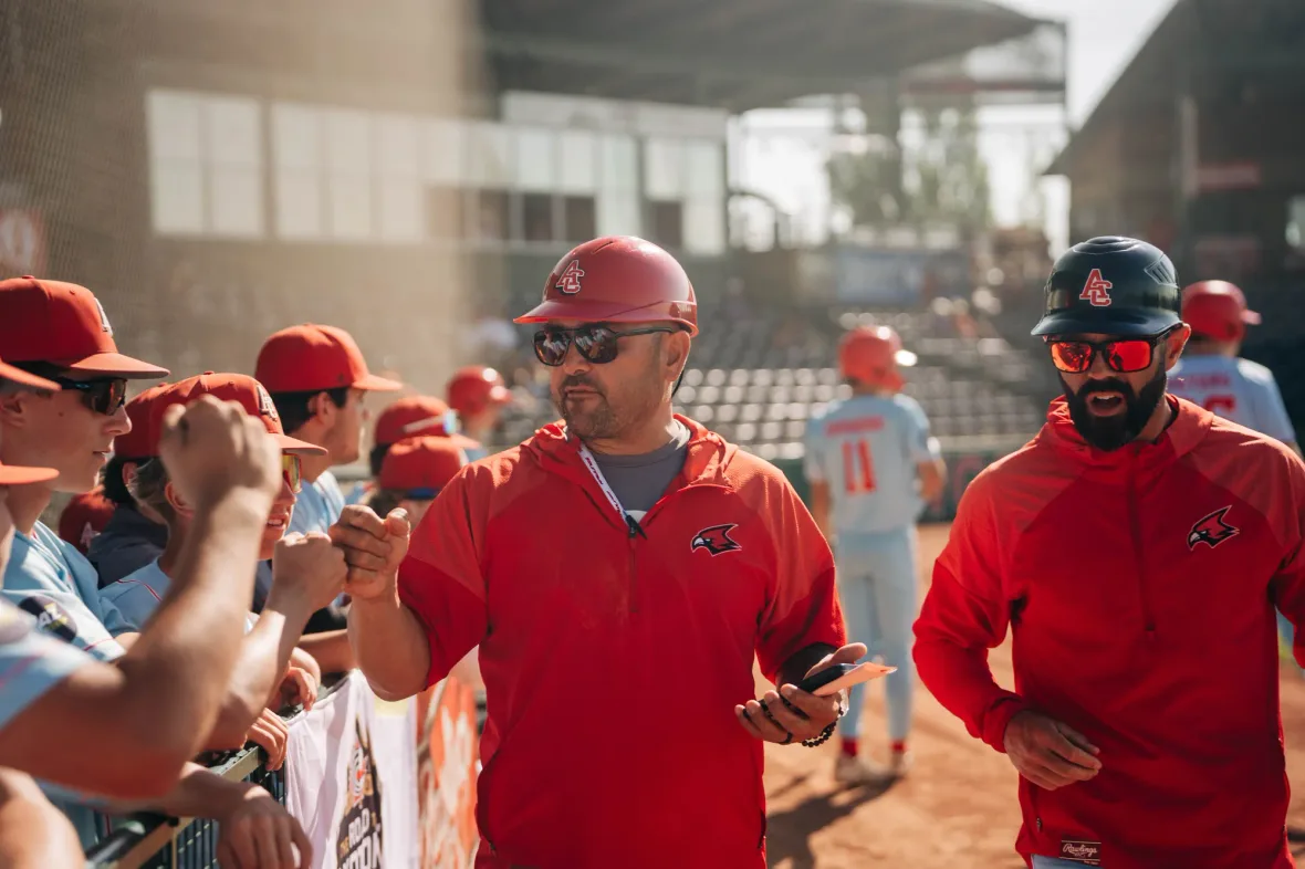 Two Cardinals coaches in red jackets and sunglasses walk along the fence at a ballpark while players in red caps reach out to fist bump.