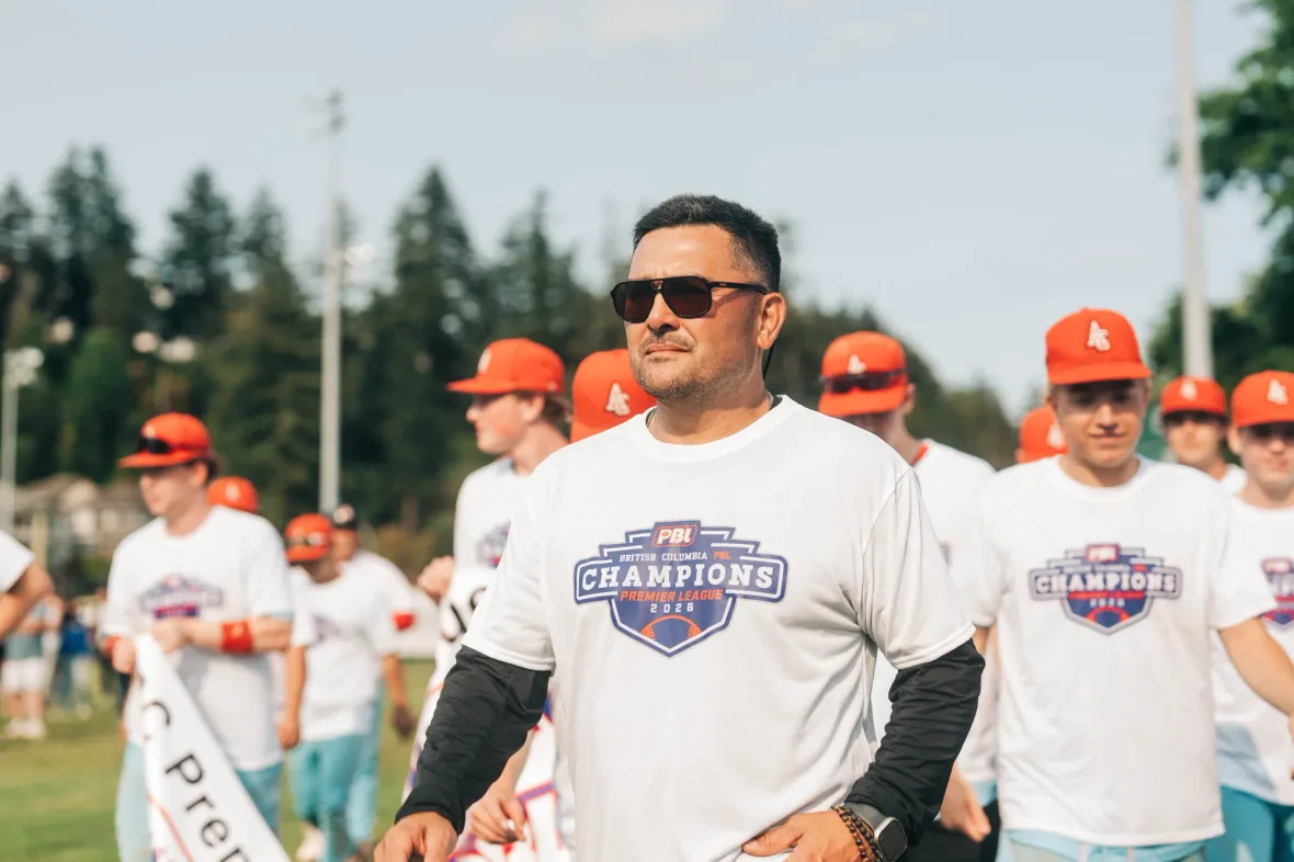 A Cardinals coach in sunglasses and a white British Columbia PBL Champions Premier League 2026 shirt walks across the field in front of players in orange caps and matching shirts.
