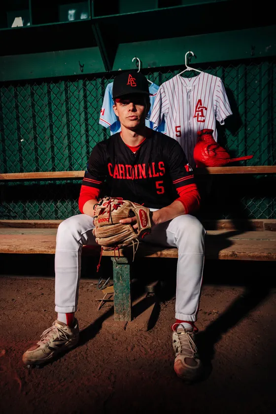 A Cardinals player sits in the dugout with the team jerseys hung behind him.